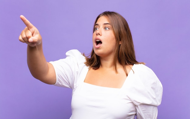 Jeune femme blonde se sentant choquée et surprise, pointant du doigt et regardant vers le haut avec admiration avec un regard étonné et bouche bée