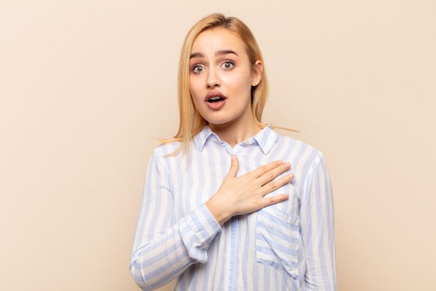 Jeune femme blonde se sentant choquée et surprise, souriante, prenant la main à cœur, heureuse d'être celle ou montrant sa gratitude