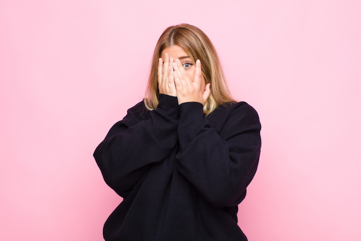 Photo jeune femme blonde se sentant effrayée ou gênée, furtivement ou espionnant avec les yeux à moitié couverts de mains sur le mur