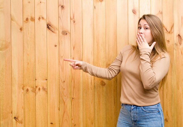 Jeune femme blonde se sentant heureuse, choquée et surprise, couvrant la bouche avec la main et pointant vers l'espace de copie latérale