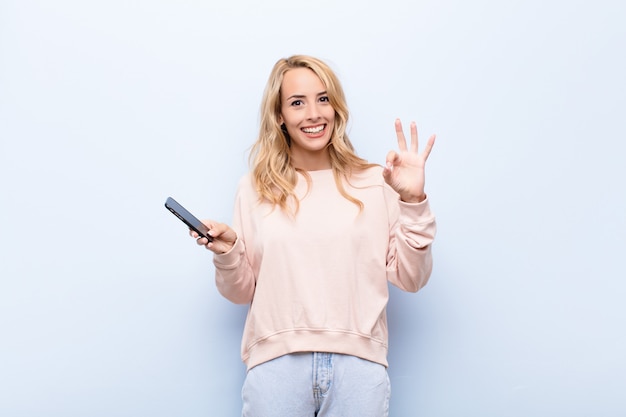 Jeune femme blonde se sentant heureuse, détendue et satisfaite, montrant son approbation avec un geste correct, souriant à l'aide d'un smartphone