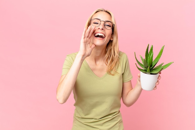 Jeune femme blonde se sentant heureuse, donnant un grand cri avec les mains à côté de la bouche et tenant un cactus