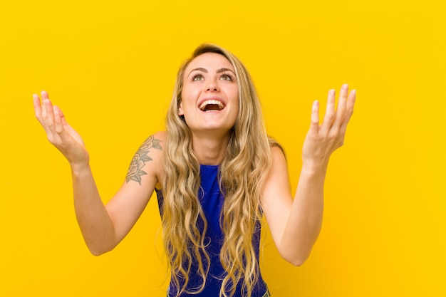 Jeune femme blonde se sentant heureuse, étonnée, chanceuse et surprise, célébrant la victoire avec les deux mains en l'air contre le mur jaune