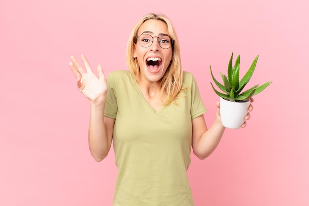 Jeune femme blonde se sentant heureuse et étonnée de quelque chose d'incroyable et tenant un cactus