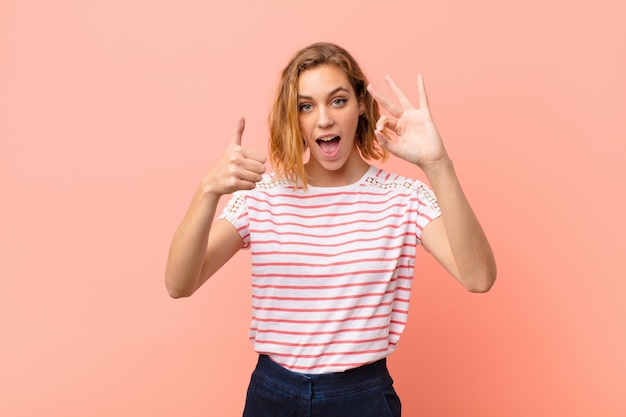 Jeune femme blonde se sentant heureuse, étonnée, satisfaite et surprise, montrant les gestes corrects et les pouces vers le haut, souriant contre le mur de couleur plat