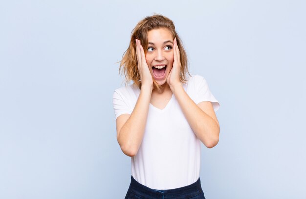 Jeune femme blonde se sentant heureuse, excitée et surprise, regardant de côté avec les deux mains sur le visage contre le mur de couleur plat