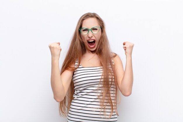 Jeune femme blonde se sentant heureuse, positive et réussie, célébrant la victoire, les réalisations ou la bonne chance contre le mur blanc