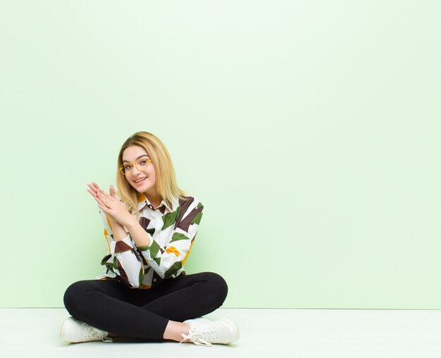 Jeune femme blonde se sentant heureuse et réussie, souriant et applaudissant, disant félicitations avec des applaudissements assis sur le sol