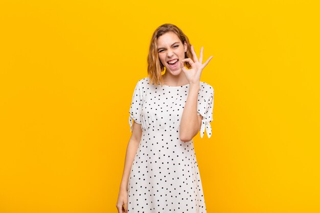 Jeune femme blonde se sentant réussie et satisfaite, souriant avec la bouche grande ouverte, faisant signe bien avec la main sur le mur de couleur plat