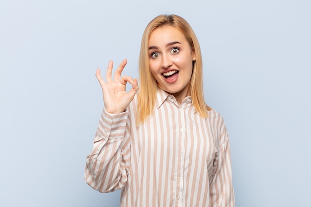 Jeune femme blonde se sentant réussie et satisfaite, souriant avec la bouche grande ouverte, faisant signe avec la main