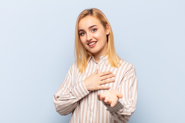 Jeune femme blonde se sentir heureuse et amoureuse, souriant d'une main à côté du cœur et de l'autre tendue à l'avant