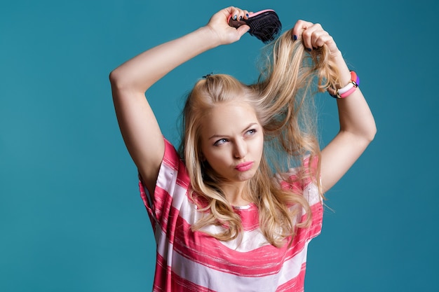 Jeune femme blonde séduisante se brosser les cheveux avec un peigne rose sur fond bleu