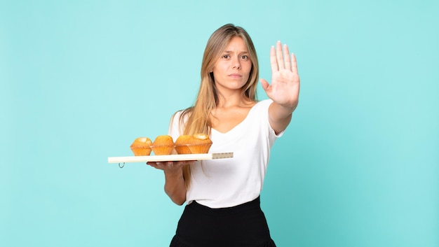 Jeune femme blonde à la sérieuse montrant la paume ouverte faisant un geste d'arrêt et tenant un muffins troy