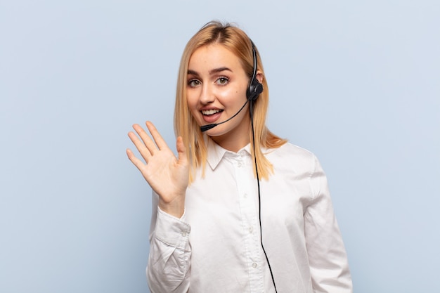 Jeune femme blonde souriant joyeusement et gaiement, agitant la main, vous accueillant et vous saluant, ou vous disant au revoir