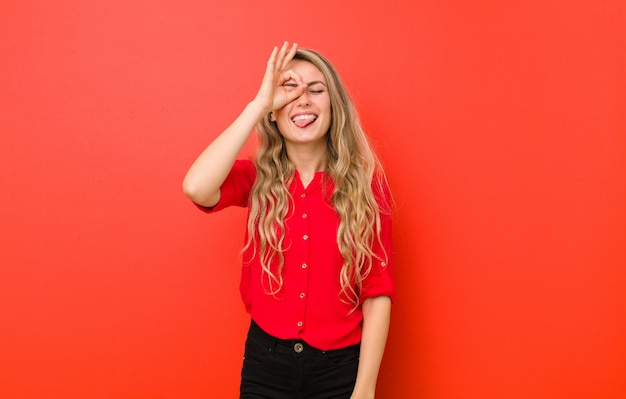 Jeune femme blonde souriant joyeusement avec grimace, plaisantant et regardant à travers un judas, espionnant des secrets sur un mur rouge