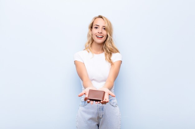 Jeune femme blonde souriant joyeusement avec un regard amical, confiant et positif, offrant et montrant un objet ou un concept tenant un portefeuille