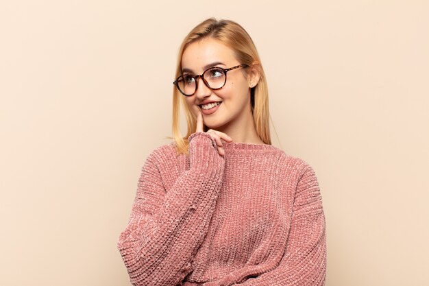 Jeune femme blonde souriant joyeusement et rêvassant ou doutant, regardant sur le côté