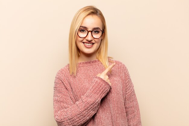 Jeune femme blonde souriant joyeusement, se sentant heureuse et pointant vers le côté et vers le haut