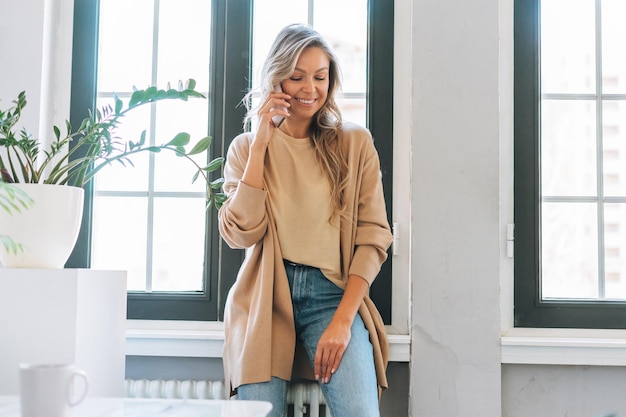 Jeune femme blonde souriante aux cheveux longs en cardigan élégant utilisant un téléphone portable dans un bureau moderne et lumineux