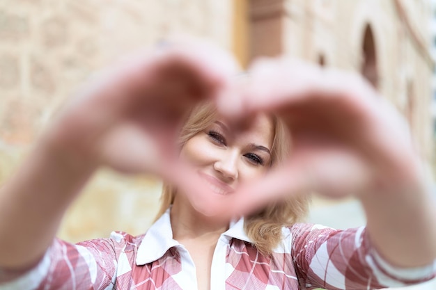 Jeune femme blonde souriante confiante faisant un geste cardiaque avec les mains dans la rue