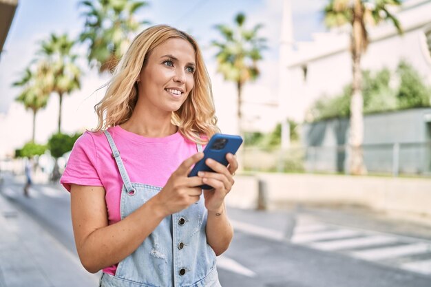 Jeune femme blonde souriante confiante utilisant un smartphone dans la rue