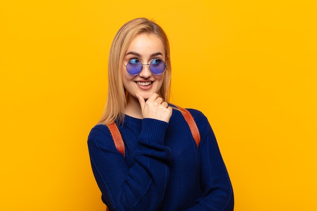 Jeune femme blonde souriante avec une expression heureuse et confiante avec la main sur le menton, se demandant et regardant sur le côté