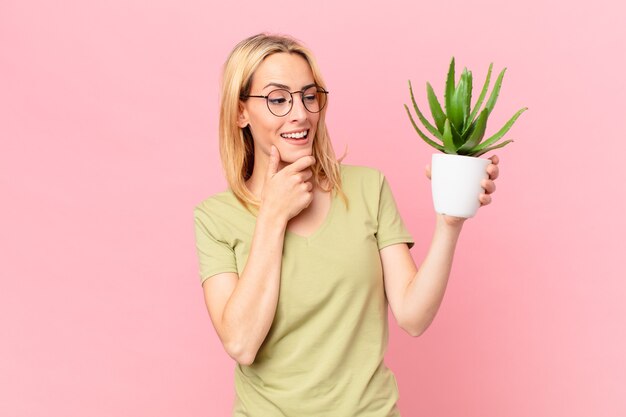 Jeune femme blonde souriante avec une expression heureuse et confiante avec la main sur le menton et tenant un cactus