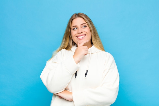 Jeune femme blonde souriante avec une expression heureuse et confiante avec la main sur le menton