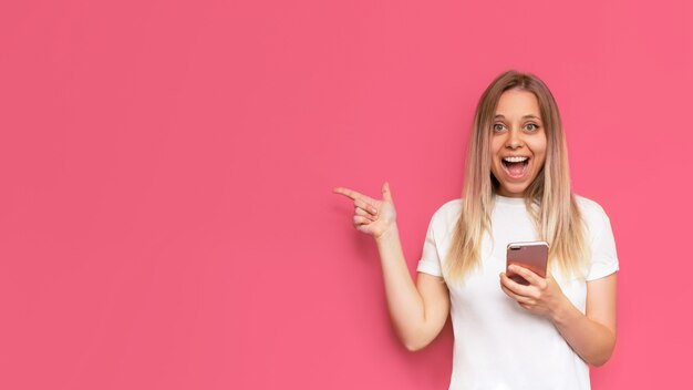 Une jeune femme blonde souriante impressionnée par un t-shirt blanc copie un espace vide pour le texte ou la conception avec son doigt tenant un téléphone portable isolé sur un mur rose de couleur vive