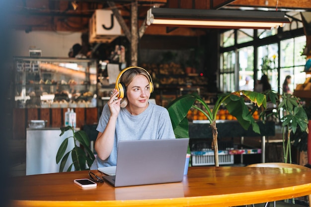 Jeune femme blonde souriante indépendante avec un casque jaune travaillant sur un ordinateur portable au café