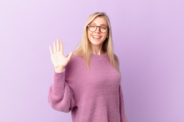 Jeune femme blonde souriante joyeusement, agitant la main, vous accueillant et vous saluant