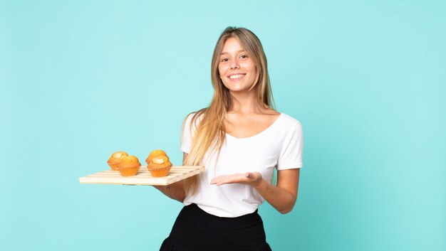 jeune femme blonde souriante joyeusement, se sentant heureuse et montrant un concept et tenant un muffins troy