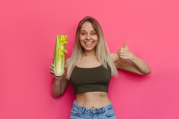 Une jeune femme blonde souriante mince caucasienne en jeans montrant le pouce vers le haut comme un geste avec sa main tenant un céleri dans sa main isolé sur un mur rose de couleur vive