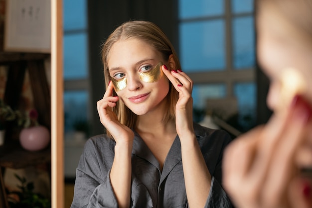 Jeune femme blonde souriante en pyjama gris soie mettant des patchs cosmétiques dorés sous les yeux tout en se tenant devant le miroir et en se regardant