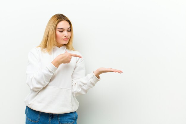 Jeune femme blonde souriante, se sentant heureuse, insouciante et satisfaite, pointant vers le concept ou l'idée sur l'espace de copie sur le côté contre le mur blanc