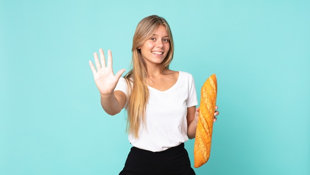 Jeune femme blonde souriante et semblant amicale, montrant le numéro cinq et tenant une baguette de pain