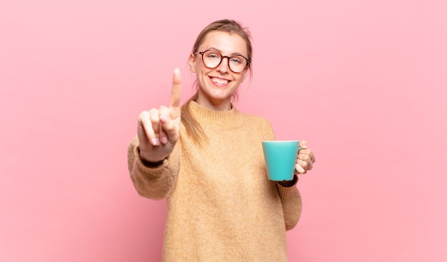 Jeune femme blonde souriante et semblant amicale, montrant le numéro un ou le premier avec la main vers l'avant, compte à rebours. concept de café