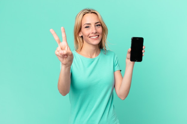 Jeune femme blonde souriante et semblant heureuse, gesticulant la victoire ou la paix et tenant un téléphone intelligent