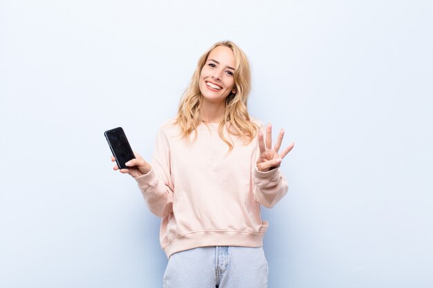 Jeune femme blonde souriante et à la sympathique, montrant le numéro quatre ou quatrième avec la main vers l'avant, compte à rebours à l'aide d'un smartphone