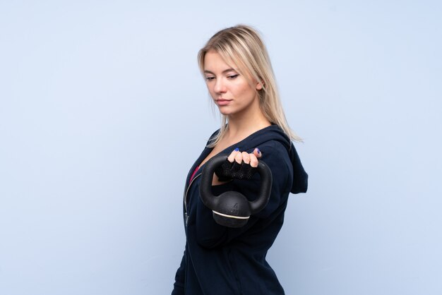 Jeune femme blonde sport sur mur bleu isolé faisant de l'haltérophilie avec kettlebell