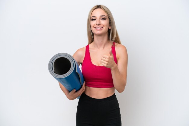 Jeune femme blonde sportive allant à des cours de yoga tout en tenant un tapis isolé sur fond blanc donnant un geste du pouce levé