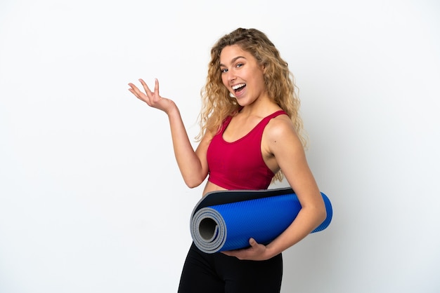 Jeune femme blonde sportive allant à des cours de yoga tout en tenant un tapis isolé sur fond blanc tendant les mains sur le côté pour inviter à venir