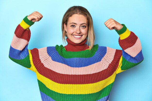 Une jeune femme blonde en sweat-shirt multicolore sur fond bleu montrant un geste de force