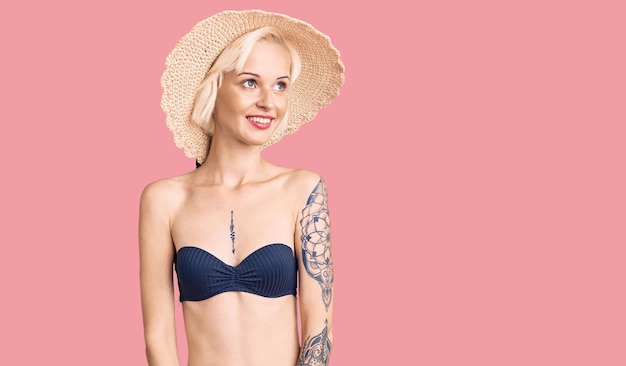 Jeune femme blonde avec un tatouage portant un bikini et un chapeau d'été regardant de côté avec un sourire sur le visage expression naturelle rire confiant