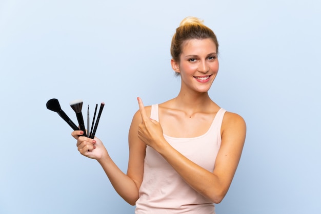 Jeune femme blonde tenant beaucoup de pinceaux de maquillage pointant sur le côté pour présenter un produit