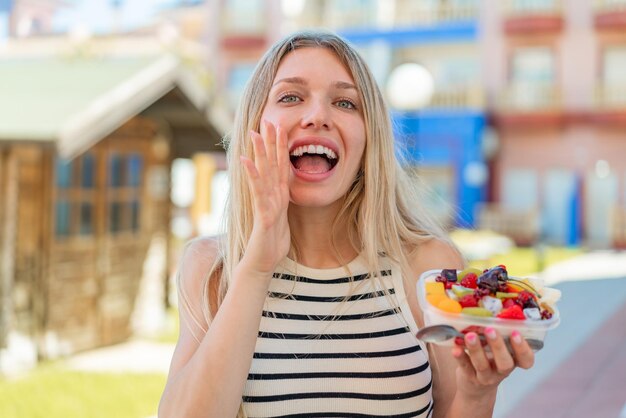 Photo jeune femme blonde tenant un bol de fruits à l'extérieur en criant avec la bouche grande ouverte