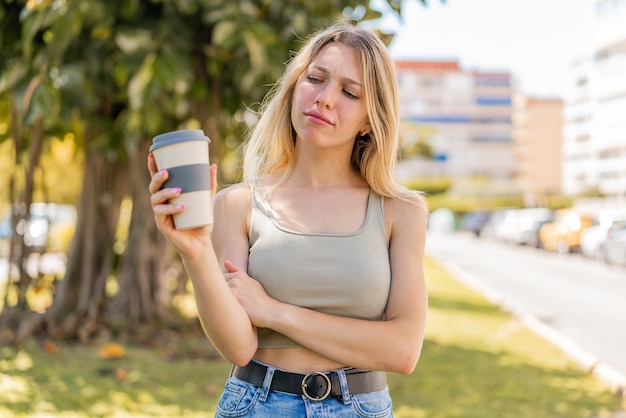 Jeune femme blonde tenant un café à emporter à l'extérieur avec une expression triste