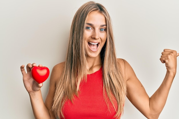 Jeune femme blonde tenant le coeur pointant le pouce vers le côté souriant heureux avec la bouche ouverte