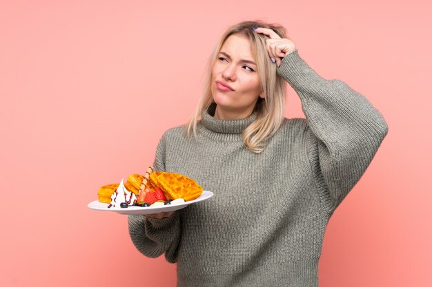 Jeune femme blonde tenant des gaufres sur un mur rose isolé ayant des doutes et avec une expression de visage confuse