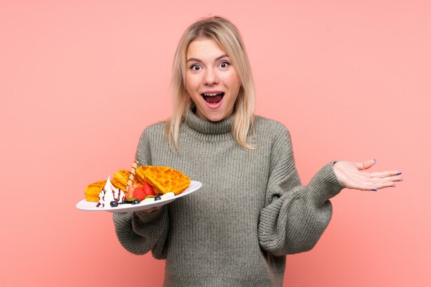 Jeune femme blonde tenant des gaufres sur un mur rose isolé avec une expression faciale choquée